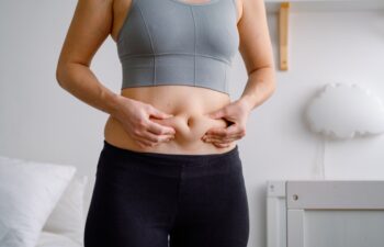 Close up of a belly with scar from c-section and abdominal fat. Women's health. A woman dressed up in sportswear demonstrating her imperfect body after a childbirth with nursery on the background.